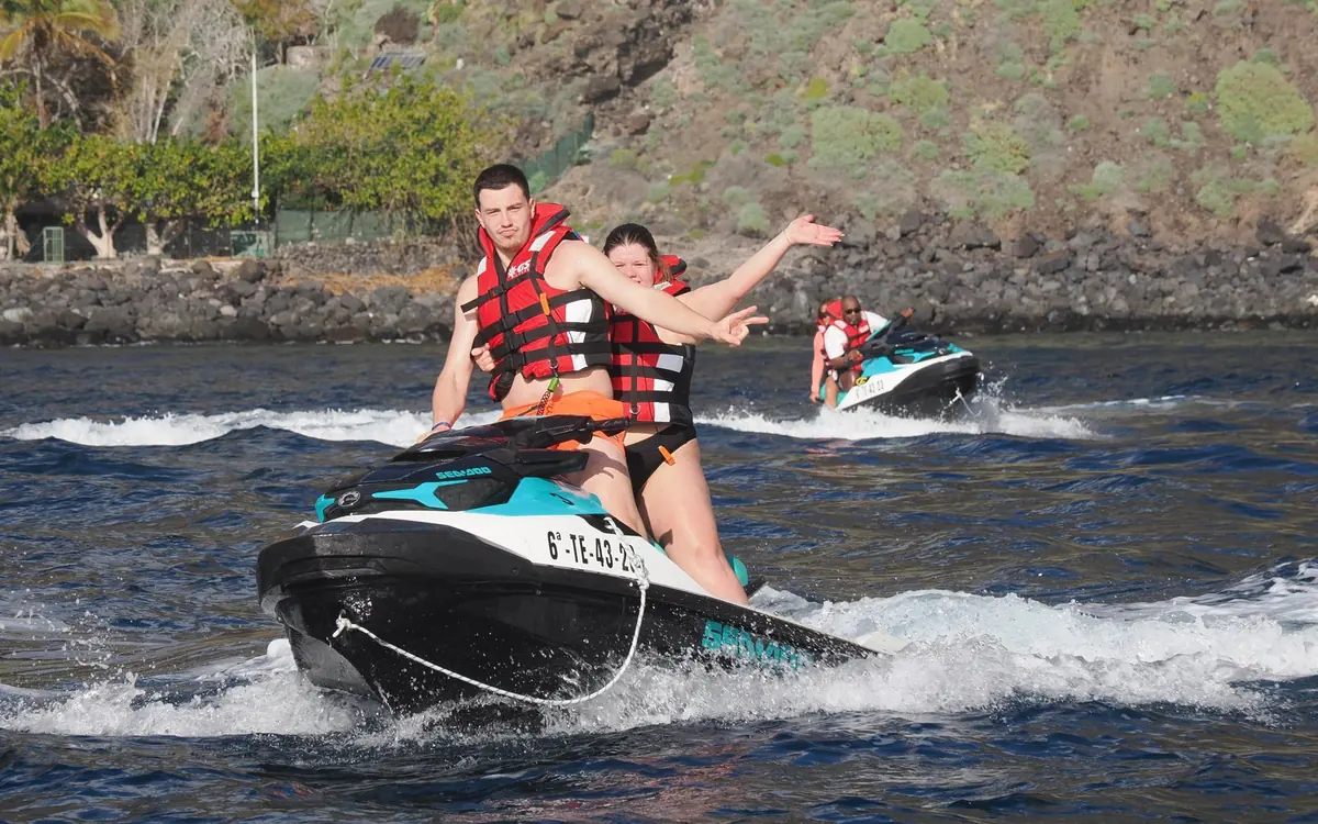 Two riders in life jackets on a jet ski, a second jet ski behind them near a rocky shore