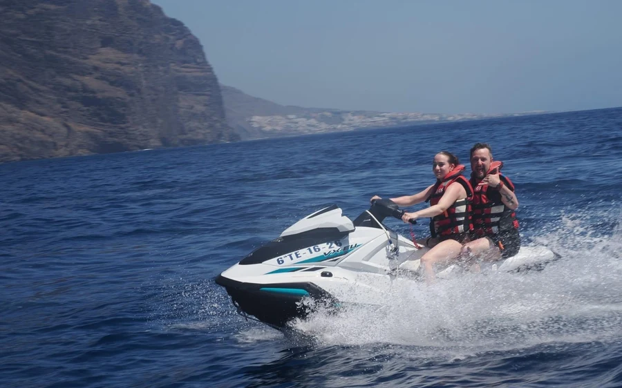 Two riders on a jet ski below tall, dark sea cliffs