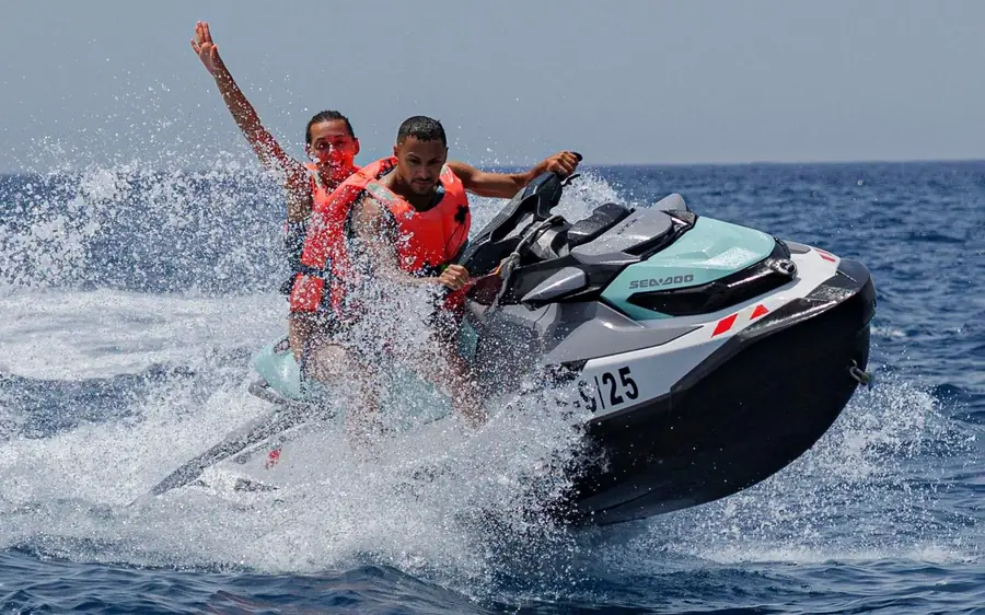 Two riders on a jet ski, one waving, in a burst of spray