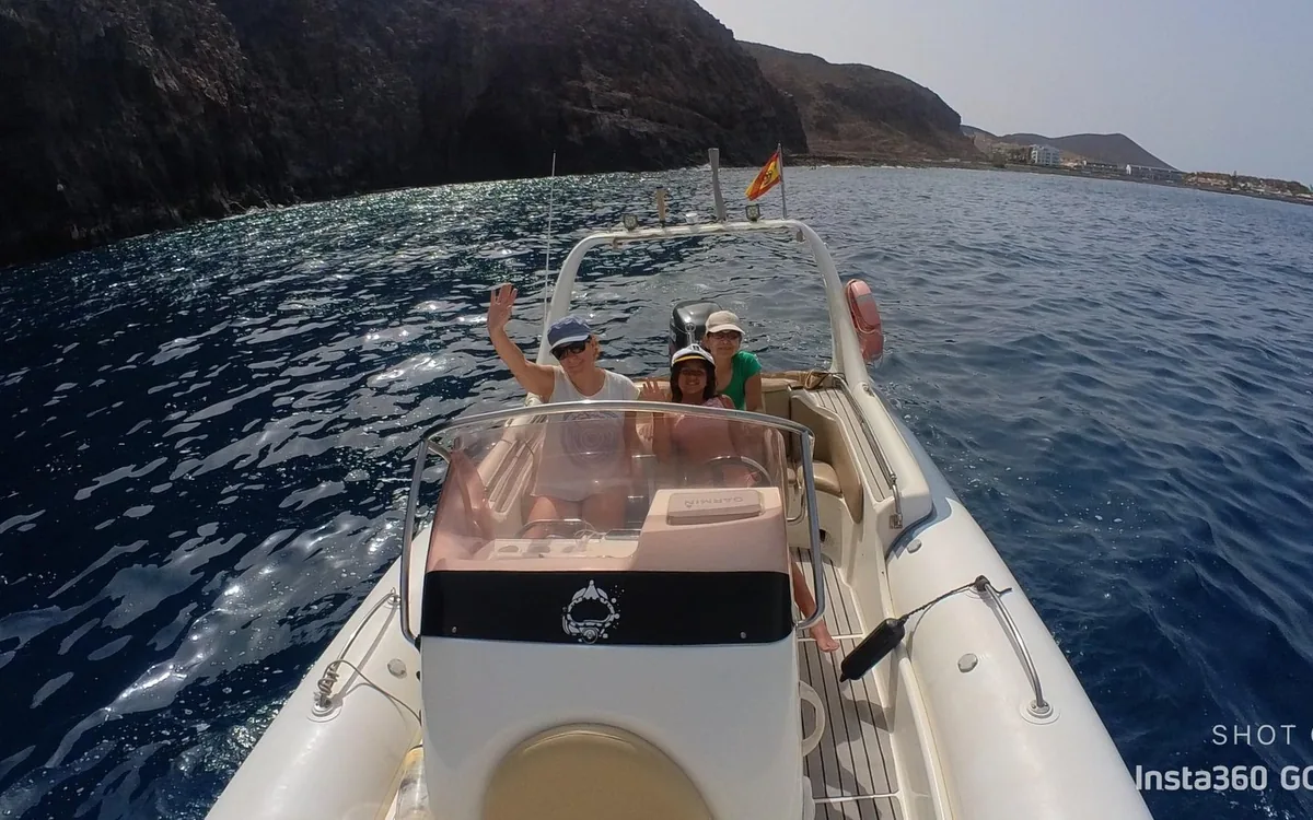 Three passengers waving from a small inflatable boat below dark coastal cliffs